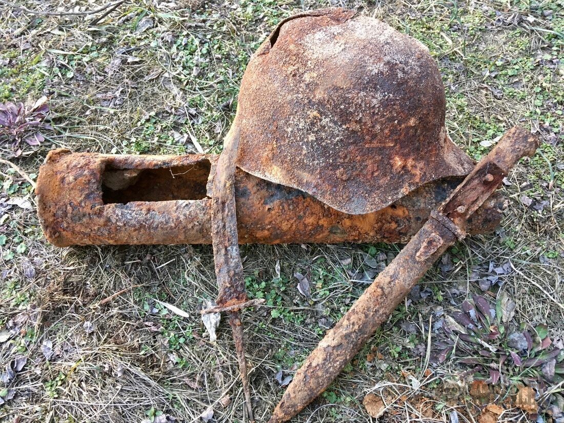 A rusted helmet shell lying on grass with a heavily corroded metal tube and two iron stakes arranged alongside it.