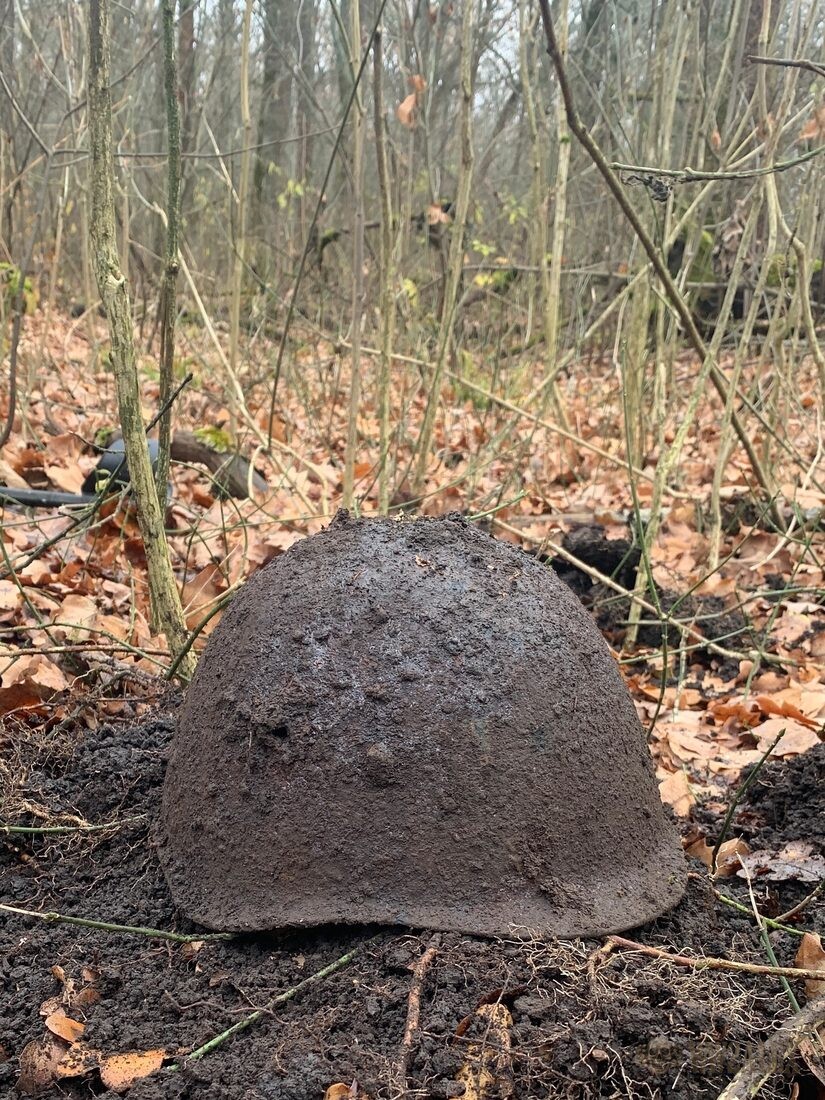 A Soviet SSH-40 helmet still caked in wet earth, resting on the ground in bare winter woodland shortly after being lifted.