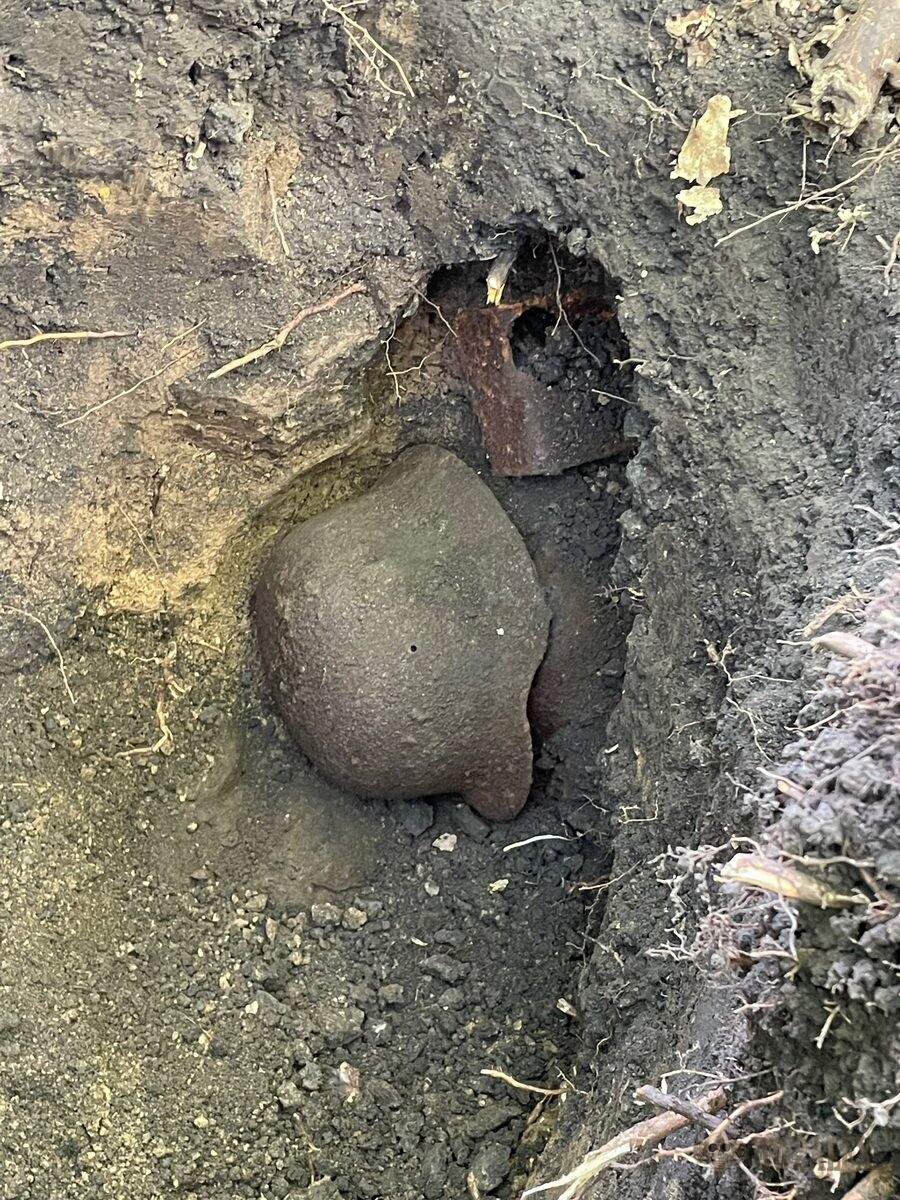 A corroded helmet shell exposed in dark soil during excavation, with a second rusted object visible behind it in the cut.