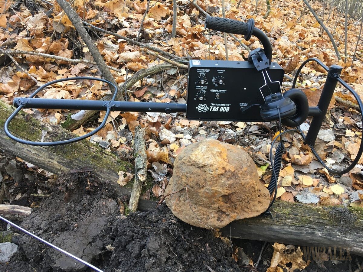 A two-box metal detector resting on a fallen log in autumn woodland, beside a heavily corroded helmet recovered at the same spot.