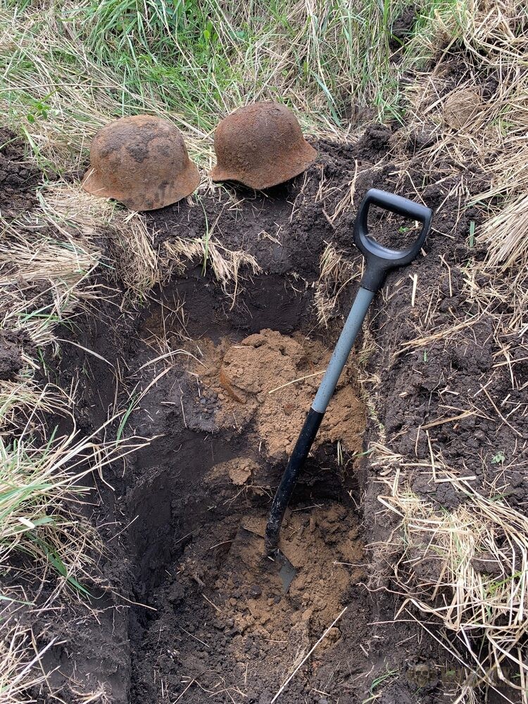 Two earth-covered helmets placed at the edge of an open excavation pit beside a spade, in a field of dry grass.